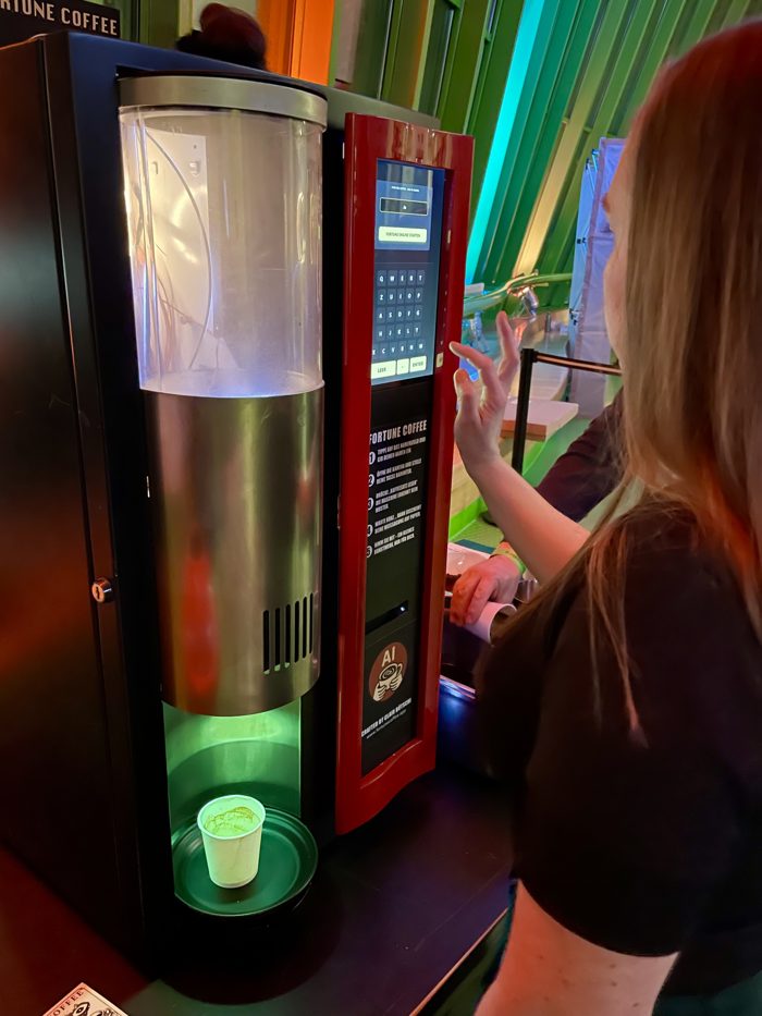 Eine Hand stellt einen Pappbecher in die Ausgabe des Automaten, dorthin, wo früher der Kaffee herauskam.